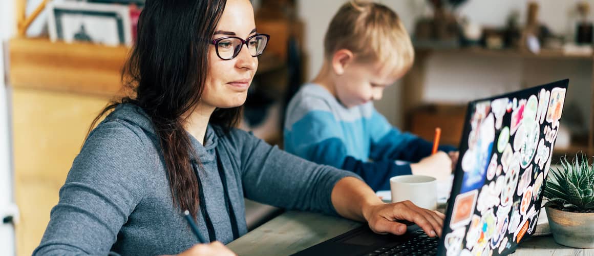 Image of woman reviewing finances on laptop at kitchen table, with son drawing next to her