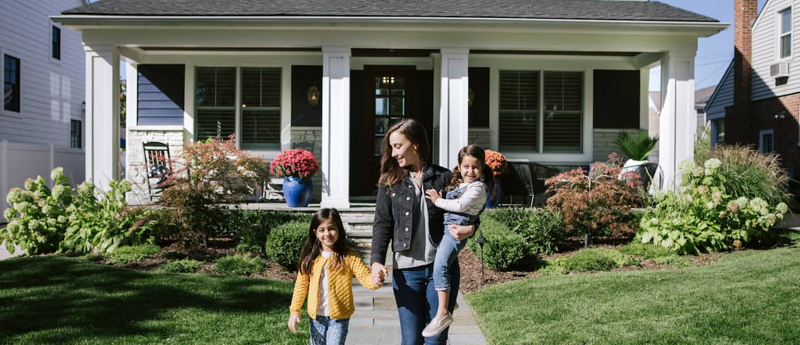 Image of mom smiling down and holding daughter's hand outside of home.