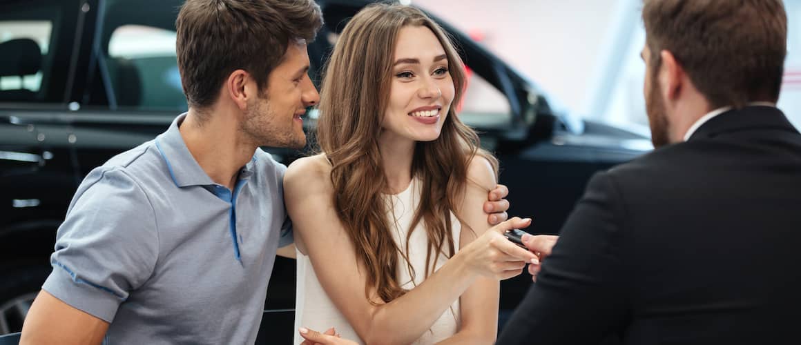 Young couple buying a car from a car dealership sales representative; shiny black car in the background.