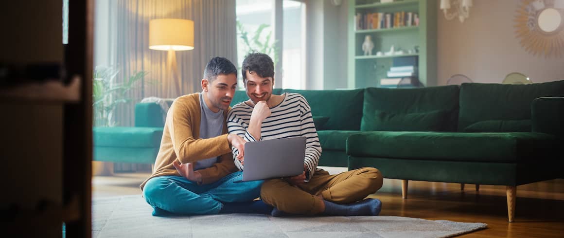 Gay couple sitting on floor of living room looking at homes available online together on laptop.