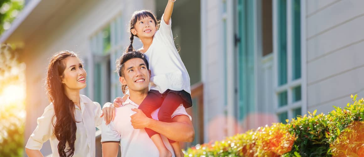 Young Asian American parents with daughter looking happy in the sunshine in front of new home.
