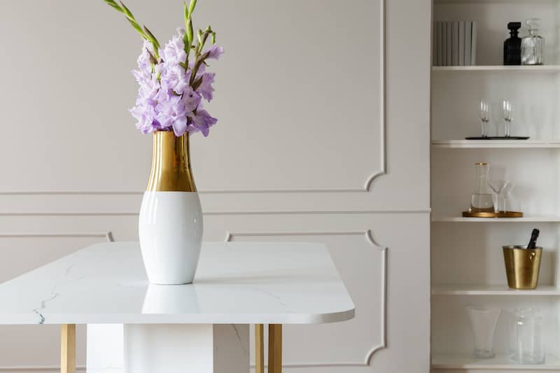 Lavender purple flowers in a golden vase on a white marble table in an elegant dining room interior with molding on beige walls.