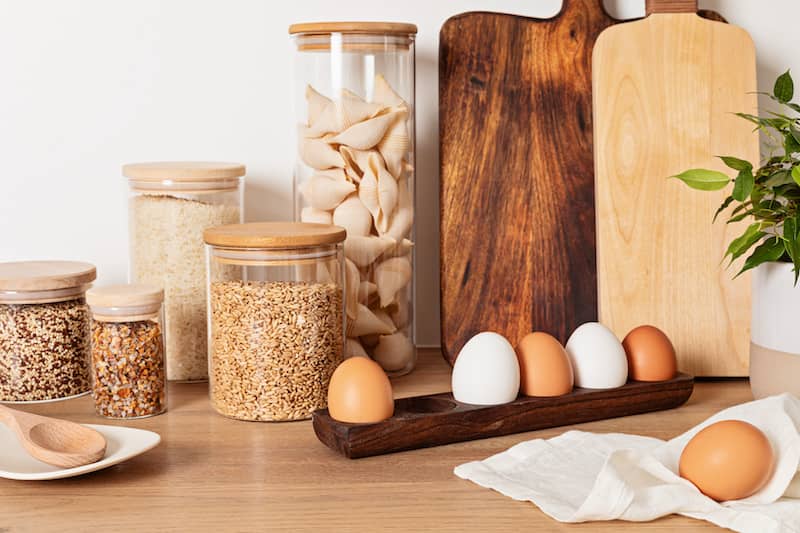 Kitchen counter with glass jars filled with grains and other food stuffs, natural wood cutting boards and farm fresh eggs in natural holder.