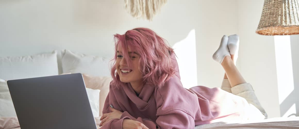 Happy hipster gen z teen girl with pink hair wear hoodie zooming on laptop lying in bed.