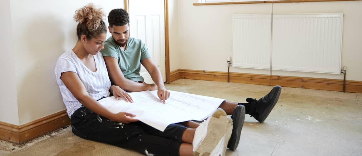 Black Couple Sitting On Floor Of Empty Room Looking At Renovation Plans
